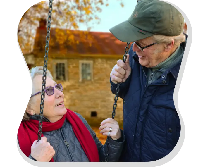 An elderly couple (man and woman) look at each other smiling while she sits on a swing. He grabs one of the swing chains while she holds both of them. In the background you can see a blurred brick house