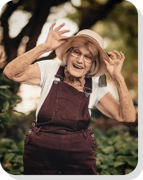 Eine alte Dame die mit beiden Händen ihre Hutkrämpe greift und glücklich in die Kamera lächelt, vor einem unscharfen Waldhintergrund